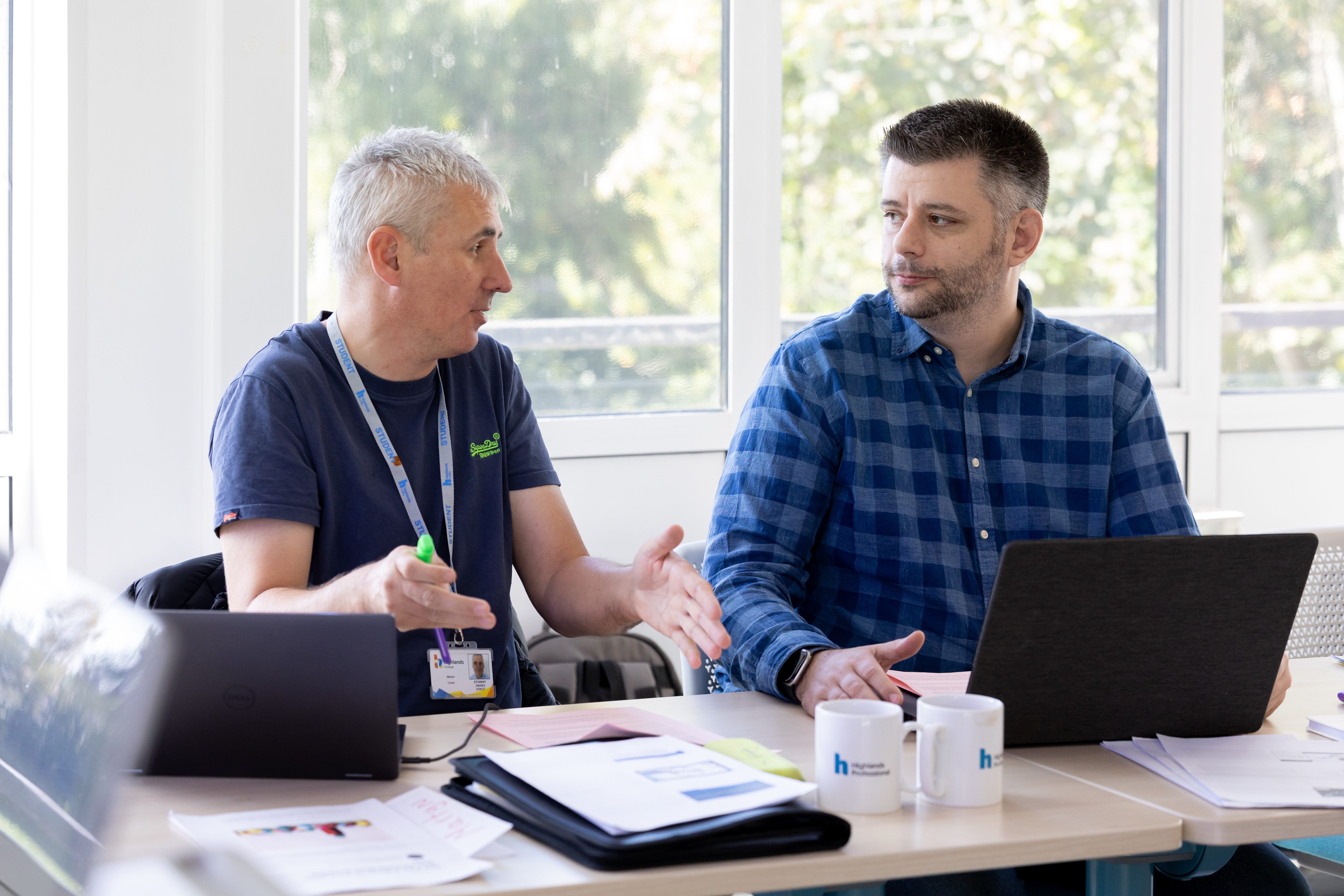 Two male adult learners sit at a table in a bright classroom, engaged in conversation while working with laptops and printed documents. One participant gestures with a marker pen while speaking, while the other looks across the table and listens. Both are wearing student lanyards. Study materials, folders, notebooks and branded mugs are arranged on the tabletop. Large windows behind the learners allow natural light into the room and provide views of greenery outside. The image depicts collaborative learning and discussion within a professional adult education setting.