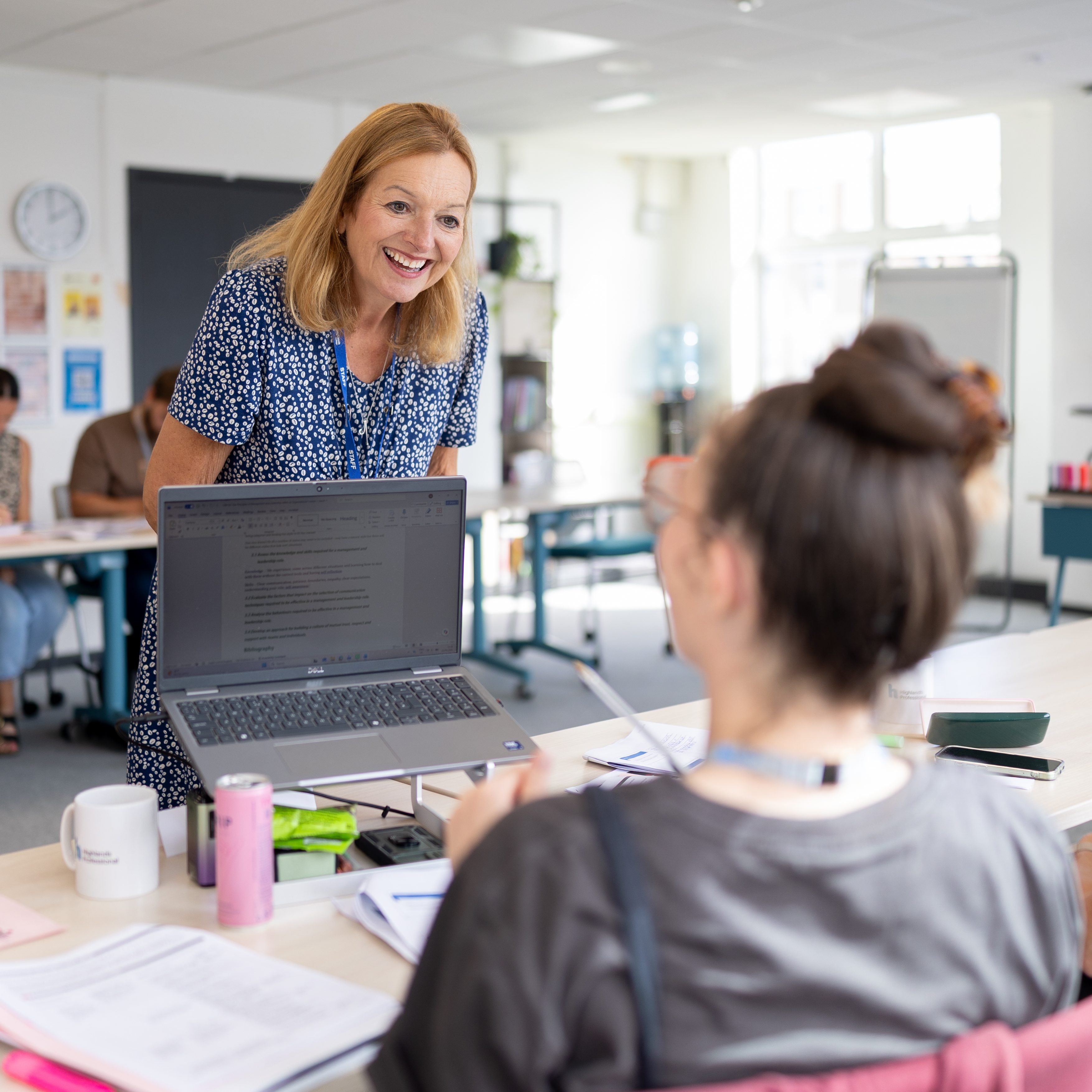 A bright, modern classroom is arranged with tables in a collaborative layout. At the centre of the image, a female tutor stands beside a table, leaning forward to engage with learners seated nearby. An open laptop on a stand faces the group, with notebooks, printed documents, mugs, pens and stationery spread across the table. Several adult learners are seated around the room, some working on laptops and others taking notes. Large windows and overhead lighting create a well-lit environment, while a presentation screen and teaching equipment are visible in the background. The image captures an active learning session focused on discussion, support and group participation.