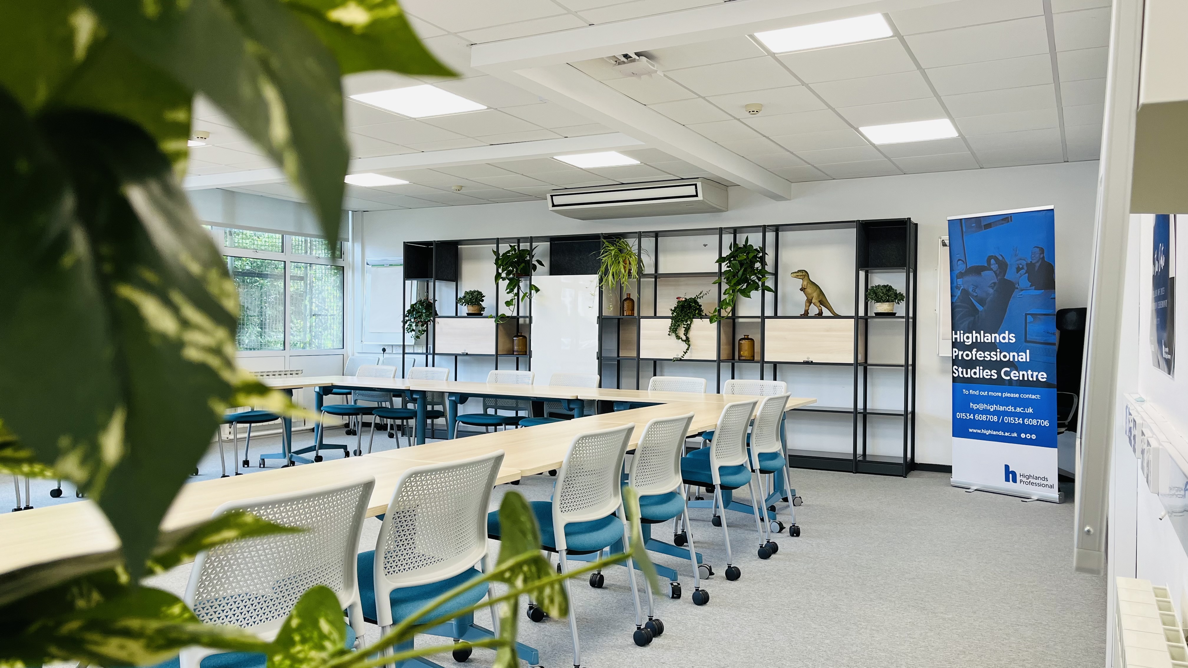Inside one of the professional classrooms, tables and chairs in blue and white, with shelving unit at the back of the room and leaves from a plant in the foreground.