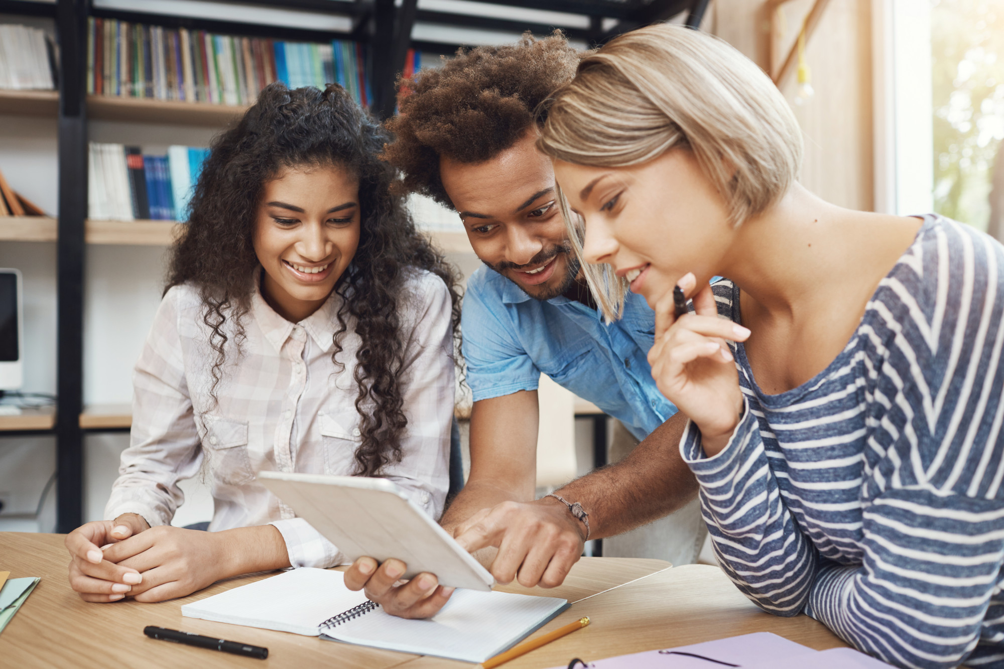 Three people stood around a table, learning on it, looking at a tablet device and smiling.