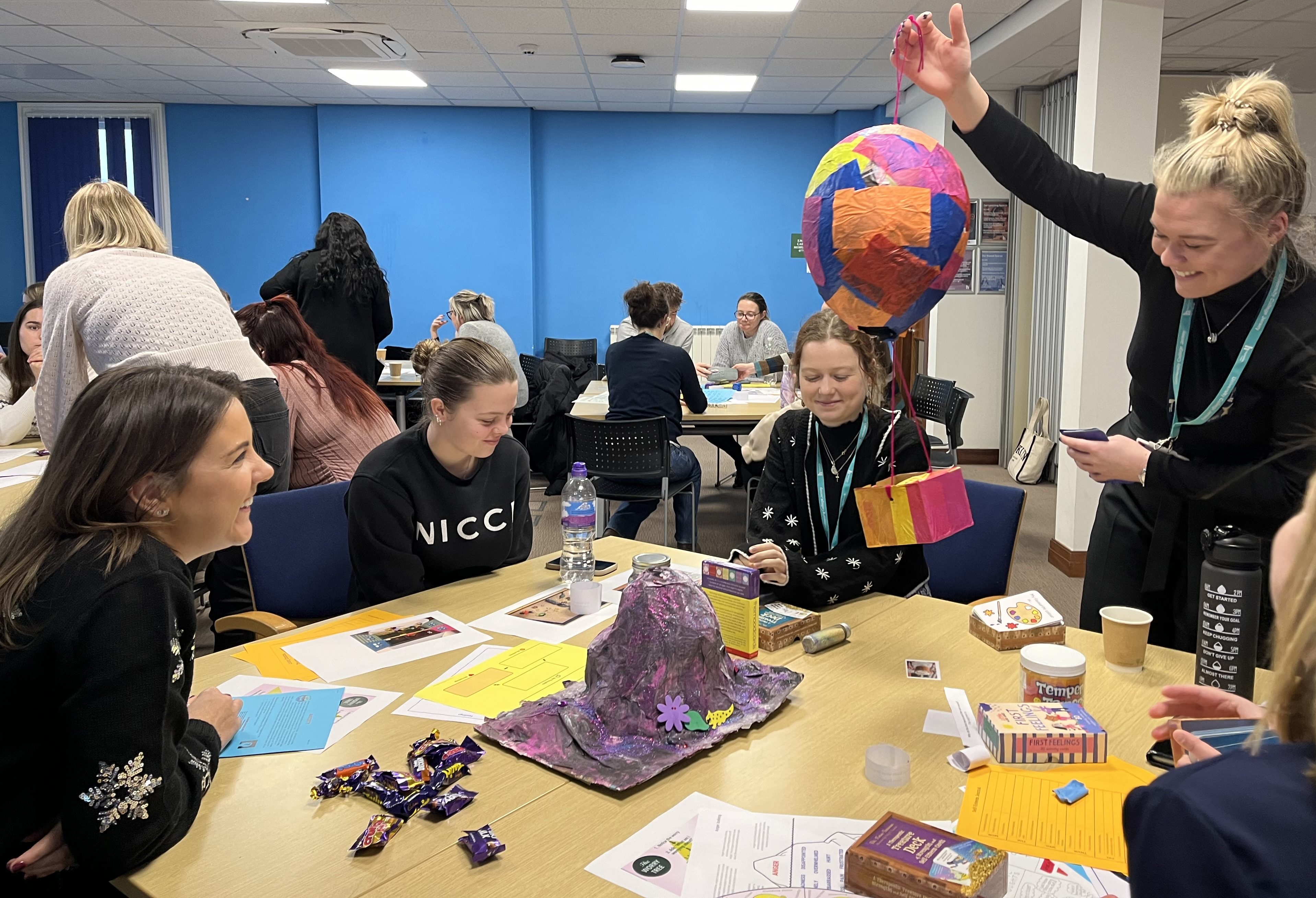 A medium shot of a classroom environment. A group sits at a table covered with chocolates, academic papers, and primary school science projects.