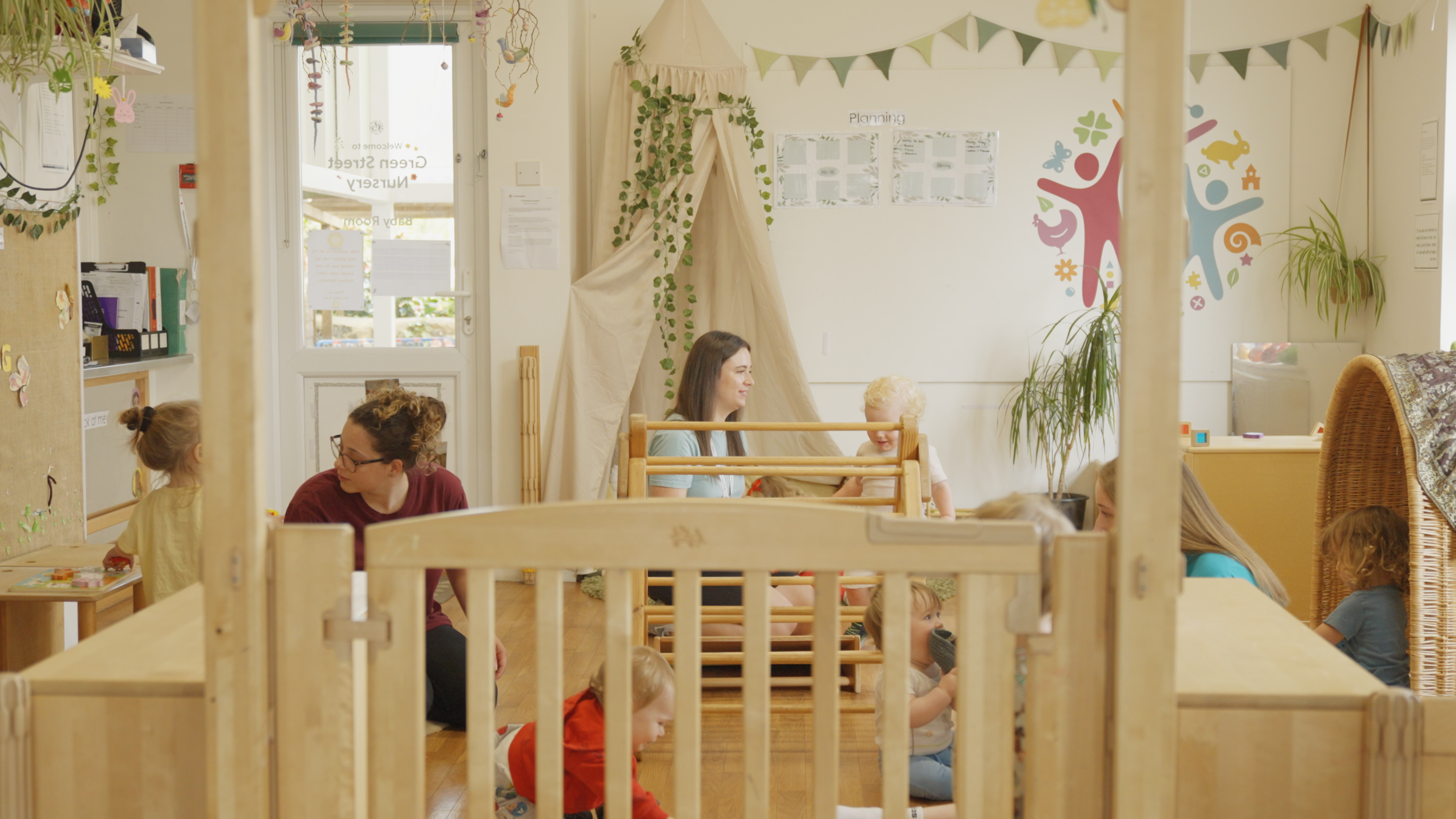 A wide shot of a playroom environment. Six babies play with toys under the supervision of three individuals.