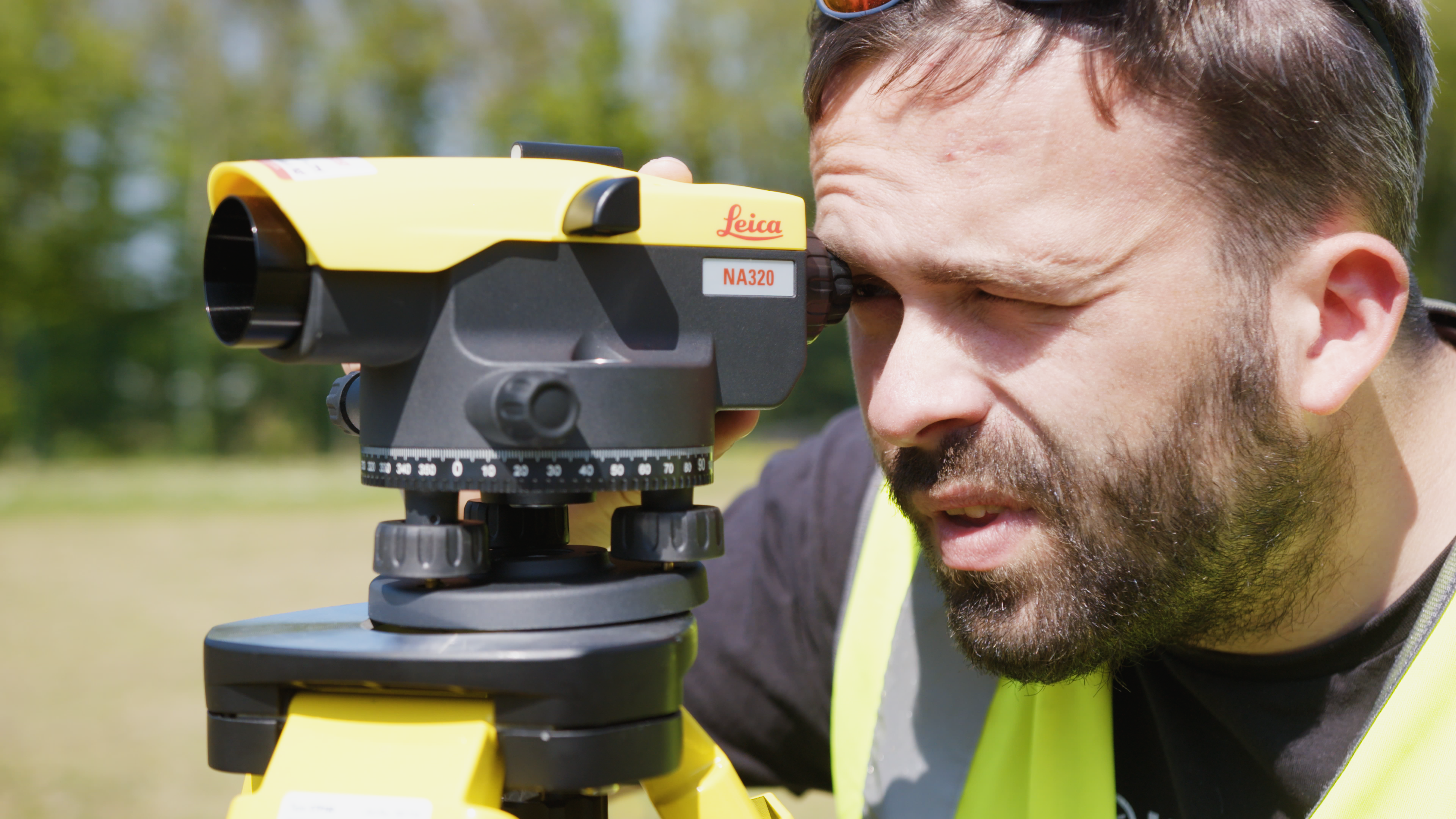 A medium close up of an individual wearing a high-visibility jacket looking through a construction measuring tool.