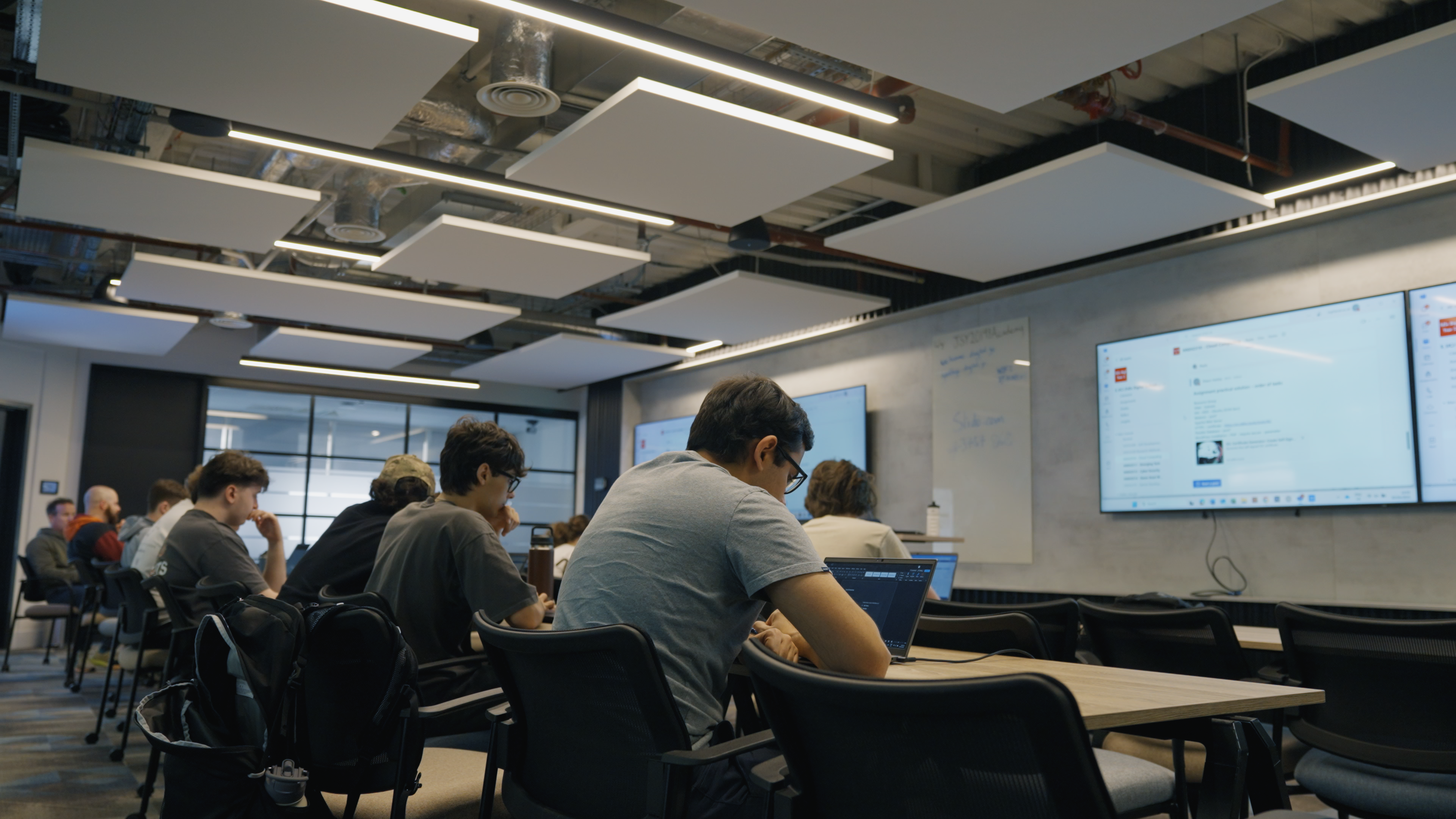 A medium wide shot of students sitting at desks in front of TV screens. There are laptops on the desks with notes on their screens.