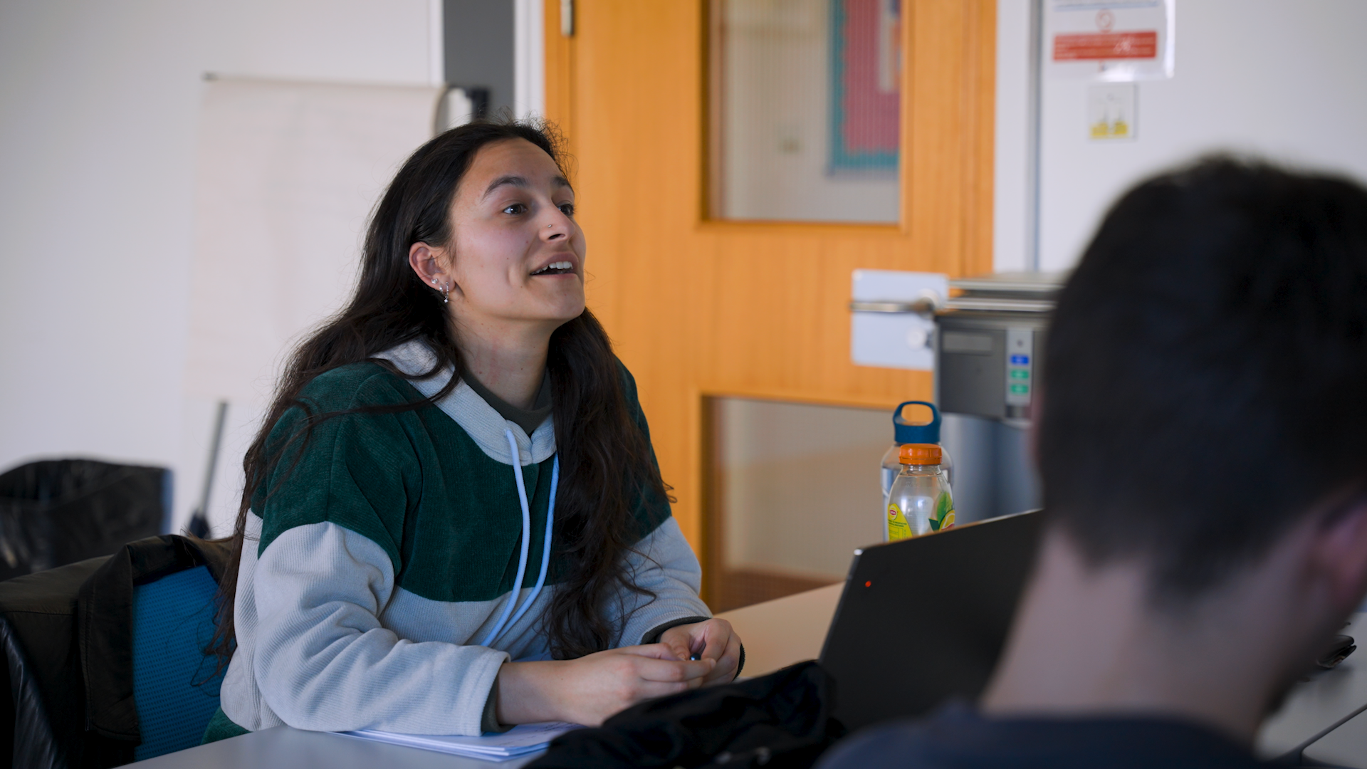 A medium shot of a classroom environment. An individual is sat at a desk with a laptop in front of them.