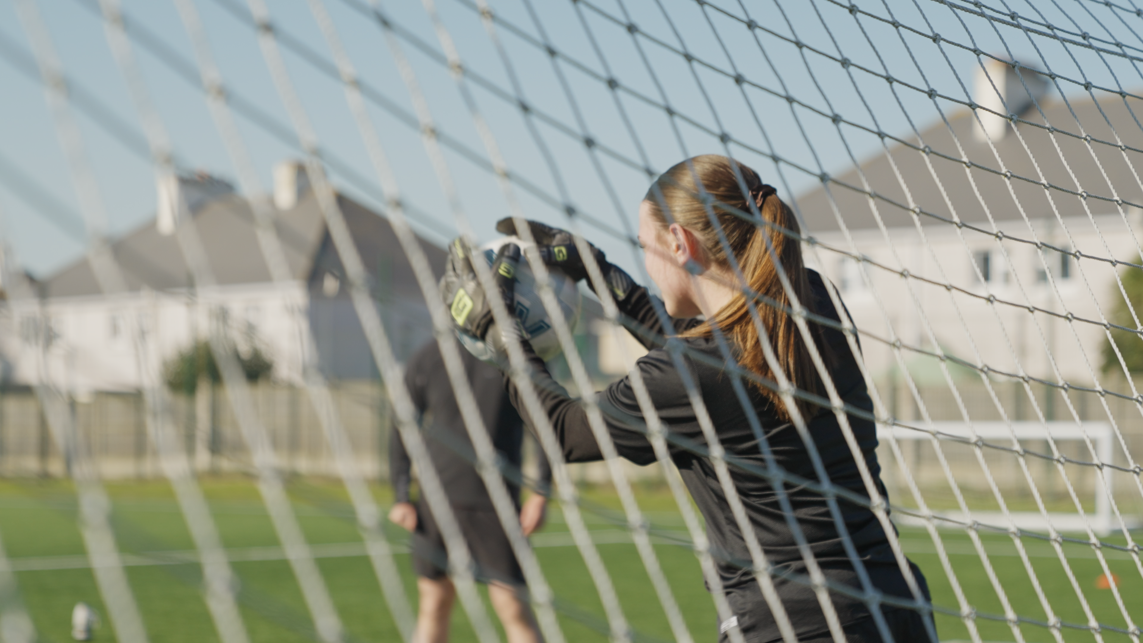 A medium shot of a goal keeper catching a football, taken from behind the goal netting. A blurred individual stands in the background but their head is obstructed by the football.