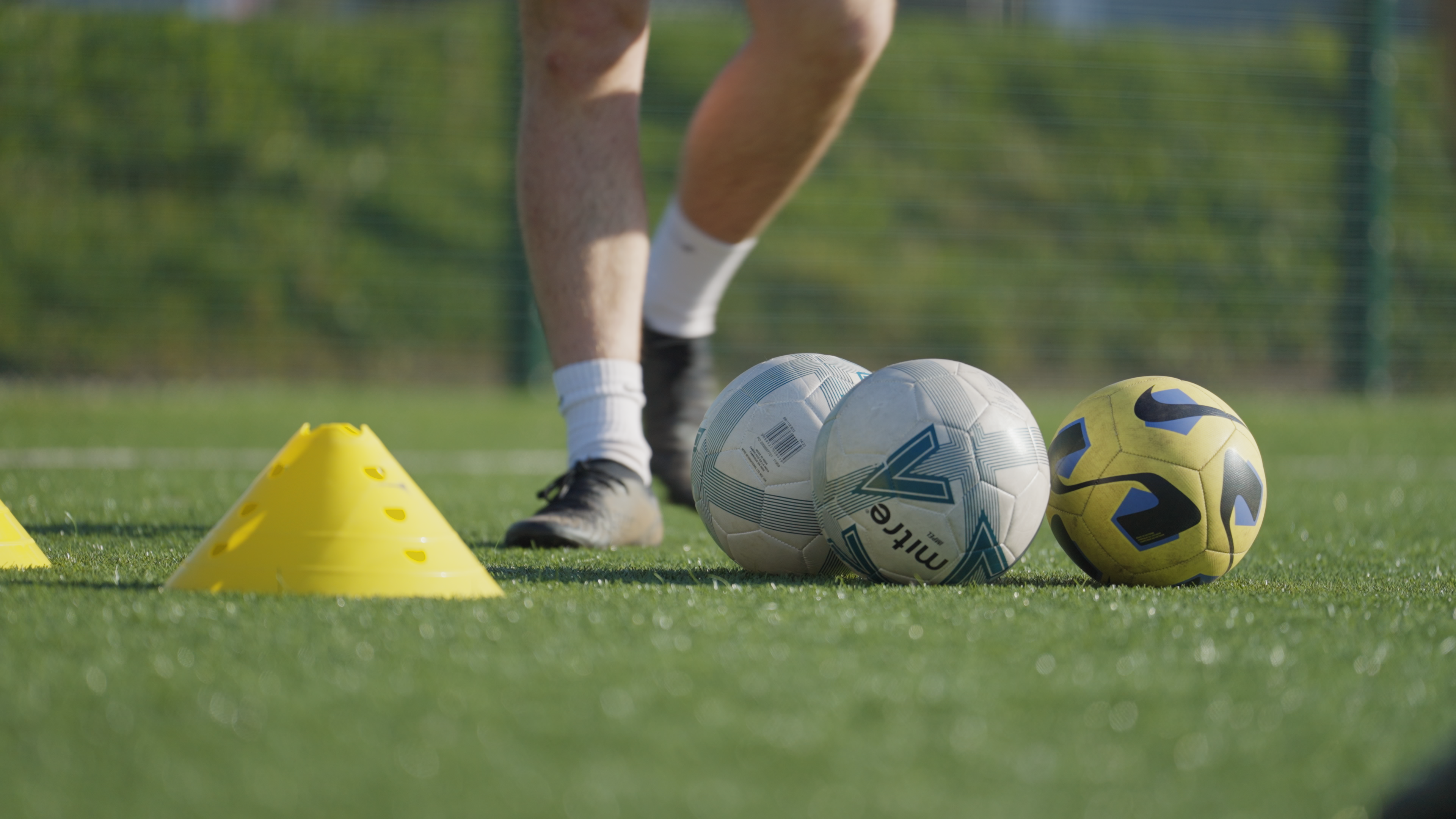 A medium shot of two legs standing on grass next to three footballs.