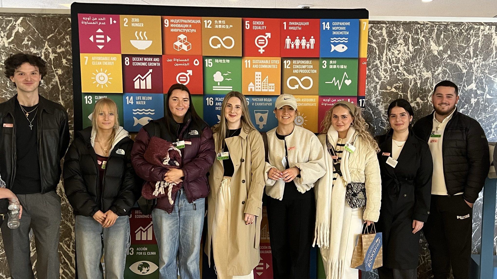A medium shot of eight individuals looking at the camera standing in front of a small board of coloured squares with several languages written on each square.
