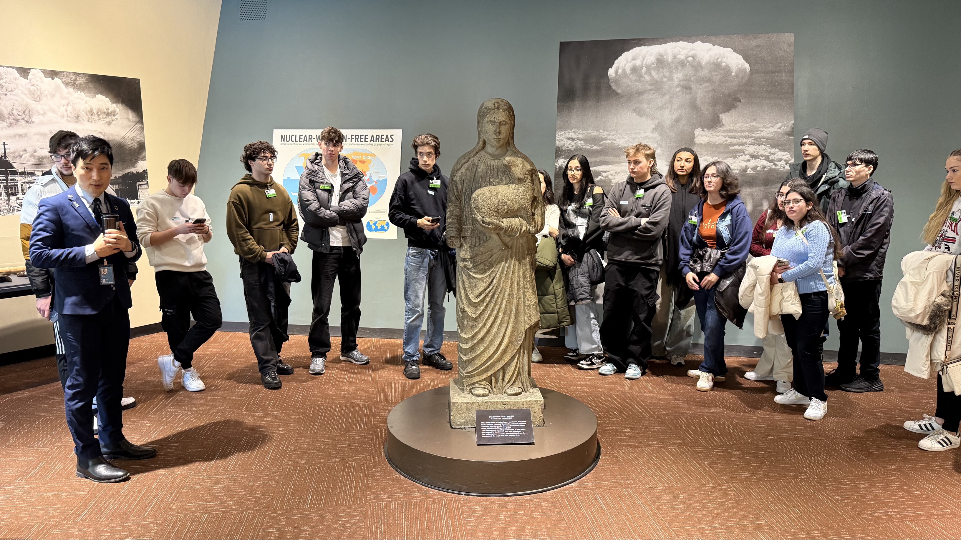 UCJ students in a room standing around a stone statue of a woman holding a lamb. On the walls behind them are photos of the first atomic bomb tests.
