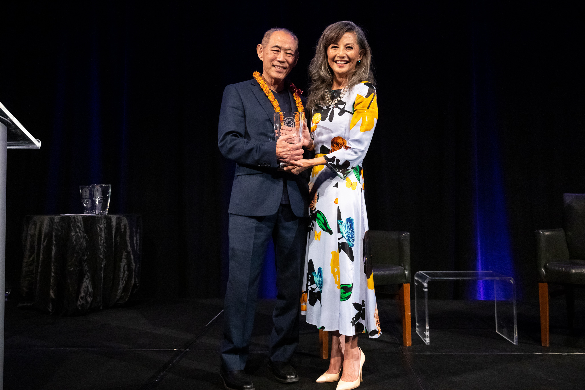 Co-Founder Dale Minami in blue suit holds award presented by Tamlyn Tomita, wearing a white and yellow dress.