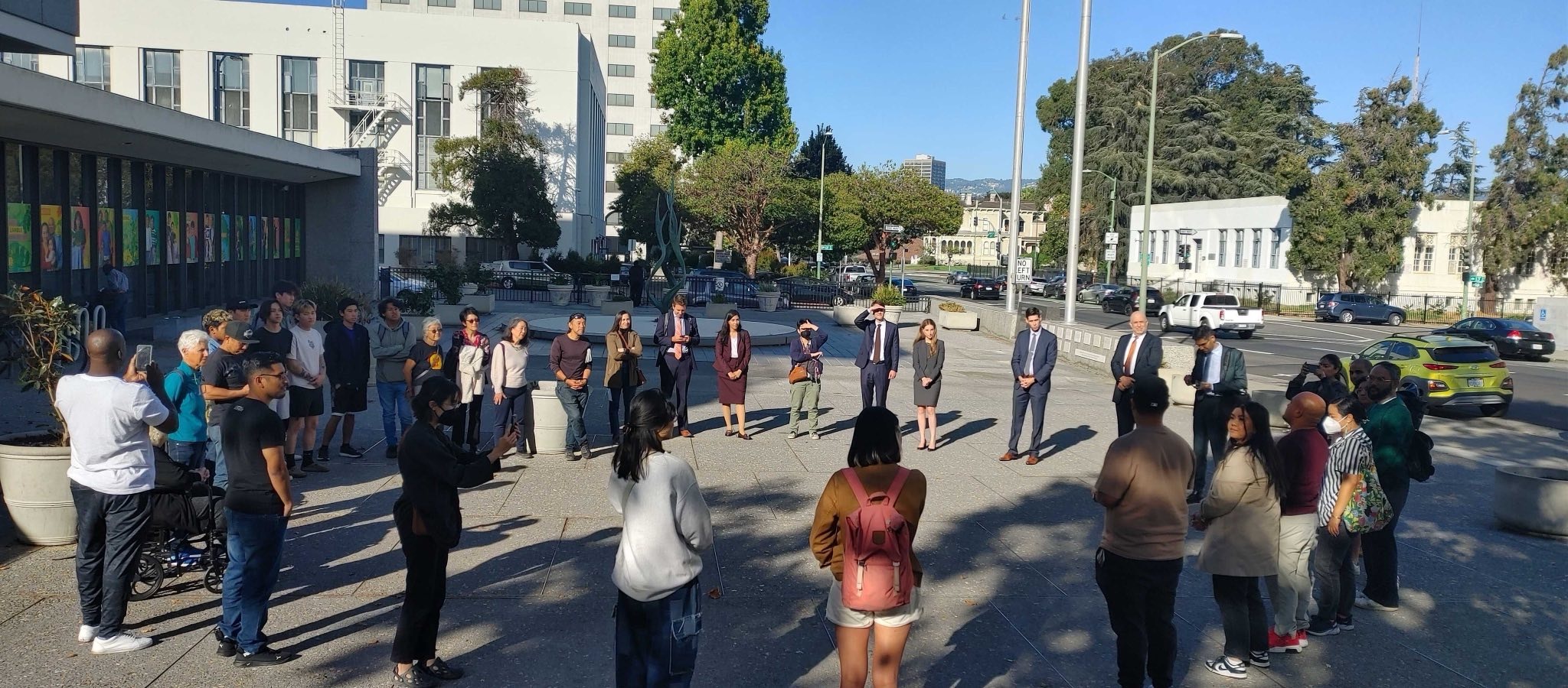 Community members, advocates, and attorneys gather outside an Oakland courthouse before a hearing in the APSC v. CDCR case. The group stands in a big circle on the plaza outside the courthouse.
