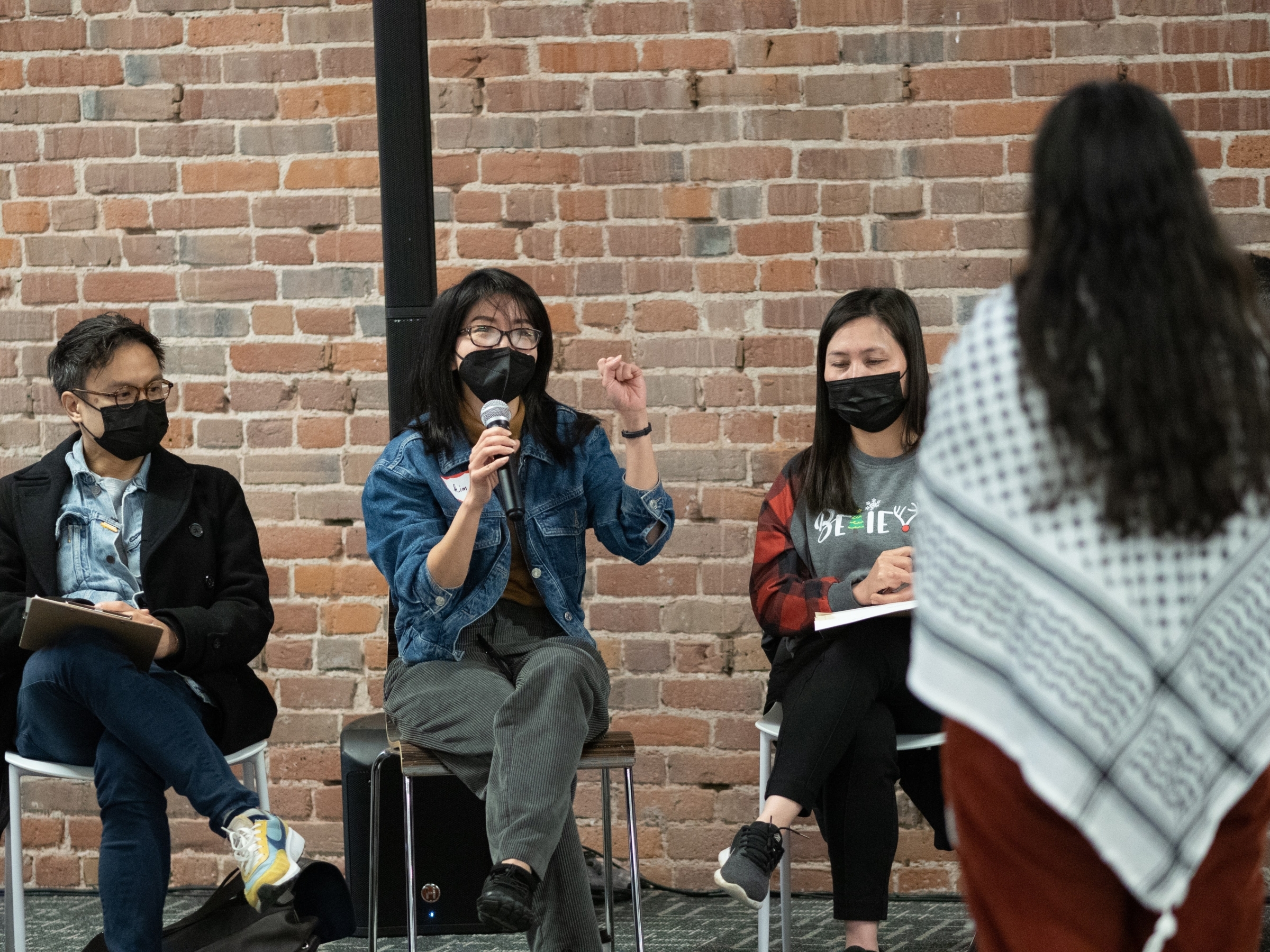 Three ALC staff members, all wearing black masks, sit on chairs in front of a brick wall. The person in the middle is holding a microphone and talking to ALC staff at our December 2023 retreat.