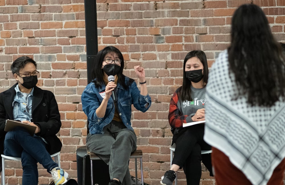 Three ALC staff members, all wearing black masks, sit on chairs in front of a brick wall. The person in the middle is holding a microphone and talking to ALC staff at our December 2023 retreat.