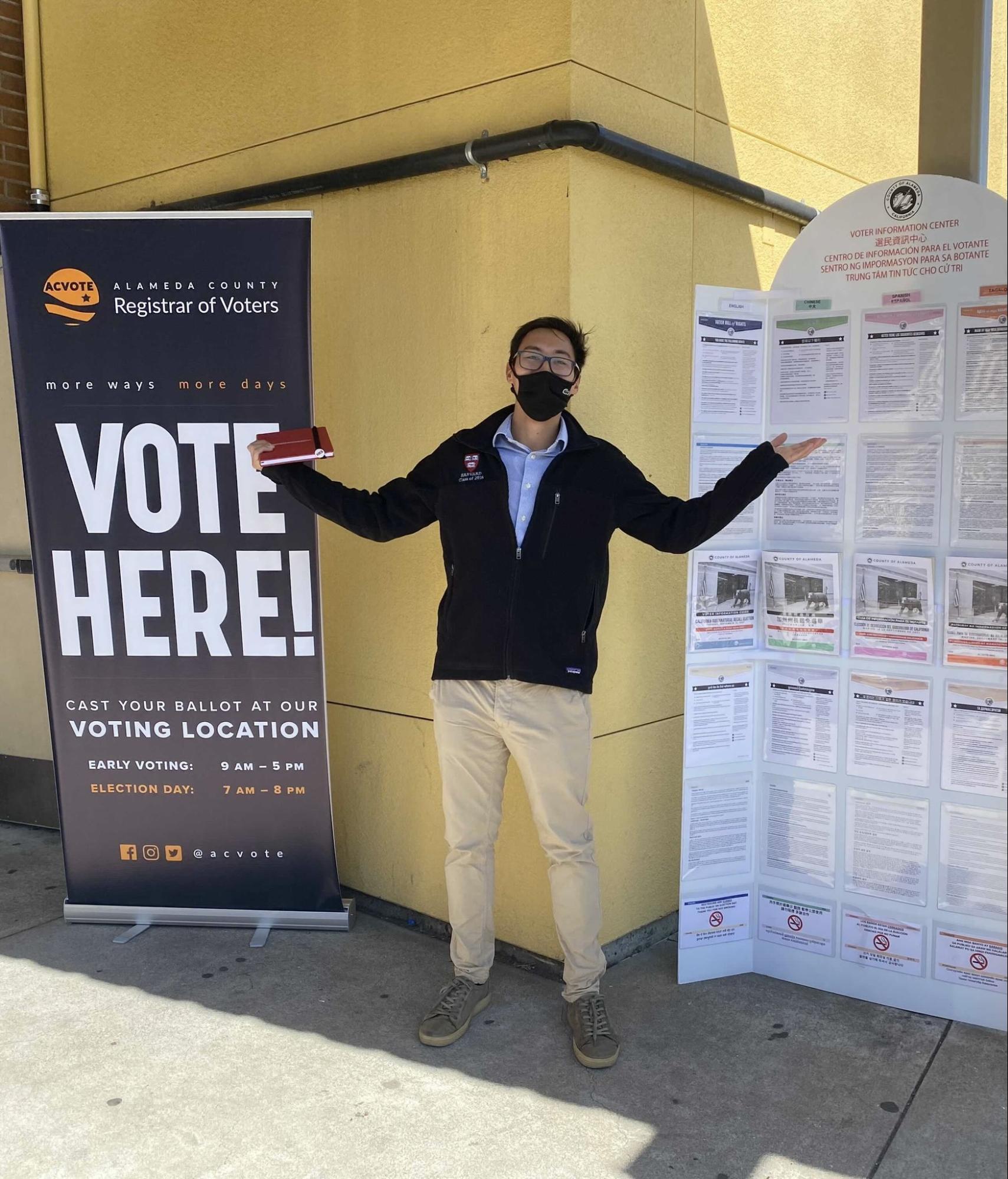 An ALC staff member stands between a "Vote Here" sign and a Voter Information sign at an Alameda voting location.