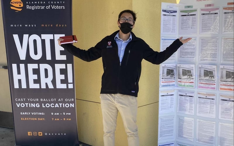 An ALC staff member stands between a "Vote Here" sign and a Voter Information sign at an Alameda voting location.