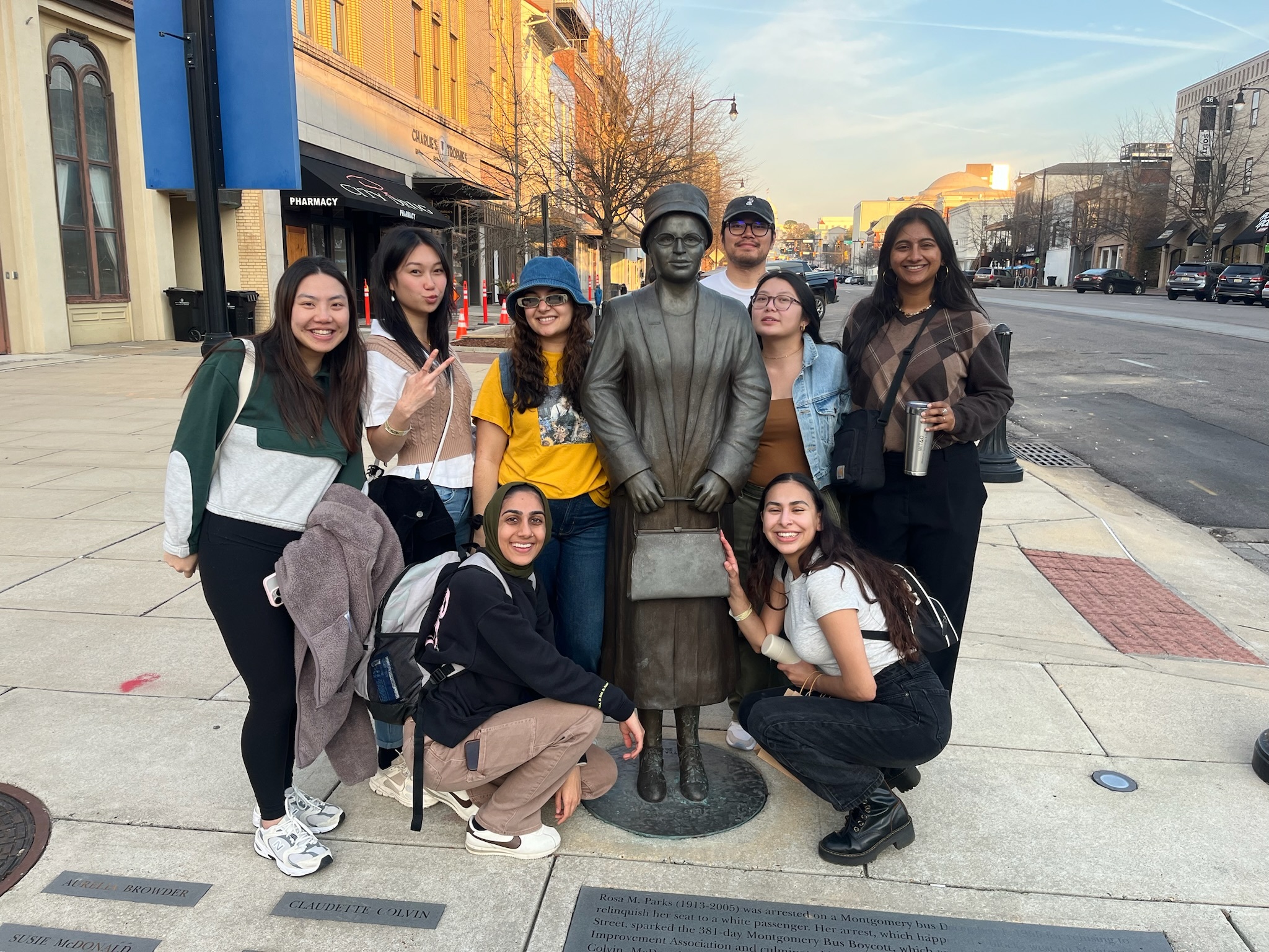 A group of 8 staff members (6 standing, 2 crouched) next to a statue of Rosa Parks at a bus stop in Montgomery, AL.