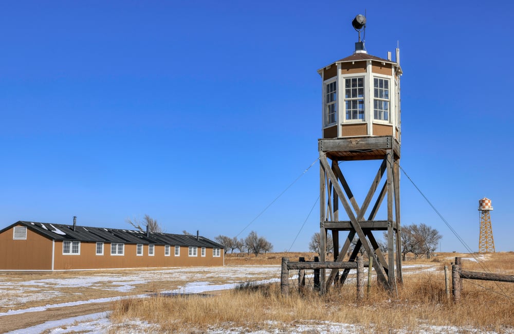 An octagonal wooden structure sits on stilts, paned windows spanning the circumference of the watch tower. Dry grass and ice stretch from the foreground to the background where a long, one-story building with several windows stands not too far from the watch tower. Much further away stands a water tower.