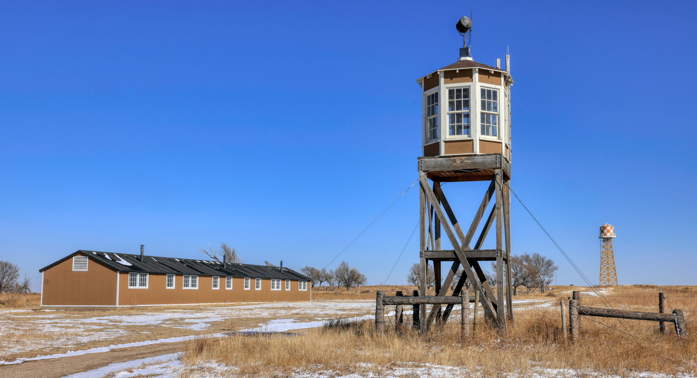 An octagonal wooden structure sits on stilts, paned windows spanning the circumference of the watch tower. Dry grass and ice stretch from the foreground to the background where a long, one-story building with several windows stands not too far from the watch tower. Much further away stands a water tower.