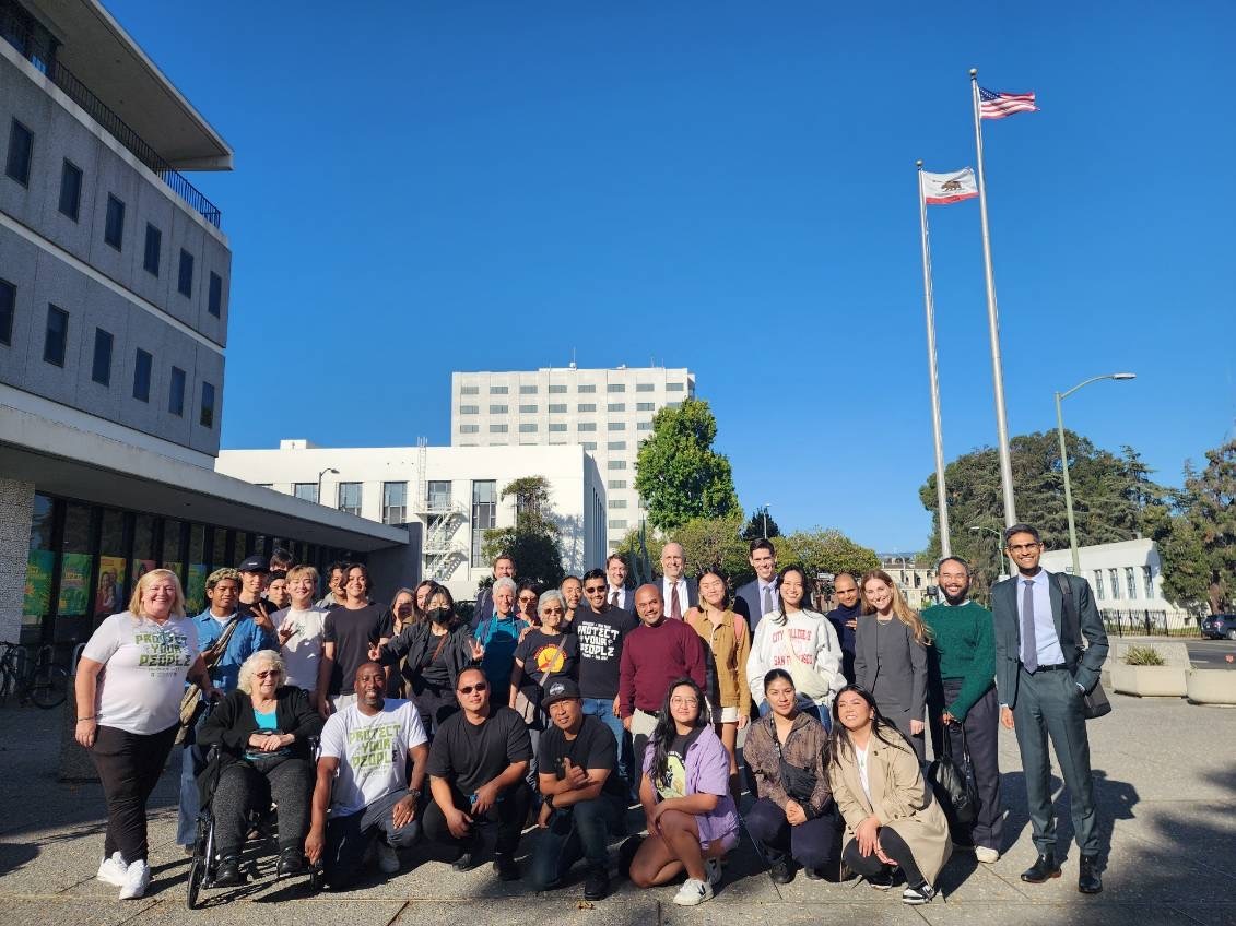 A large crowd poses for a photo outside the Alameda County Civil Courthouse. They are arranged in two rows and behind them are buildings and flagpoles bearing the US and California state flags.