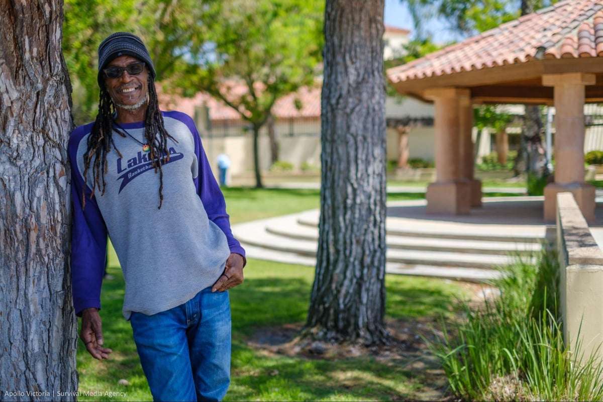 Brain Bukle smiles while leaning against a tree in a community park. He wears a gray cap and a t-shirt with purple sleeves.