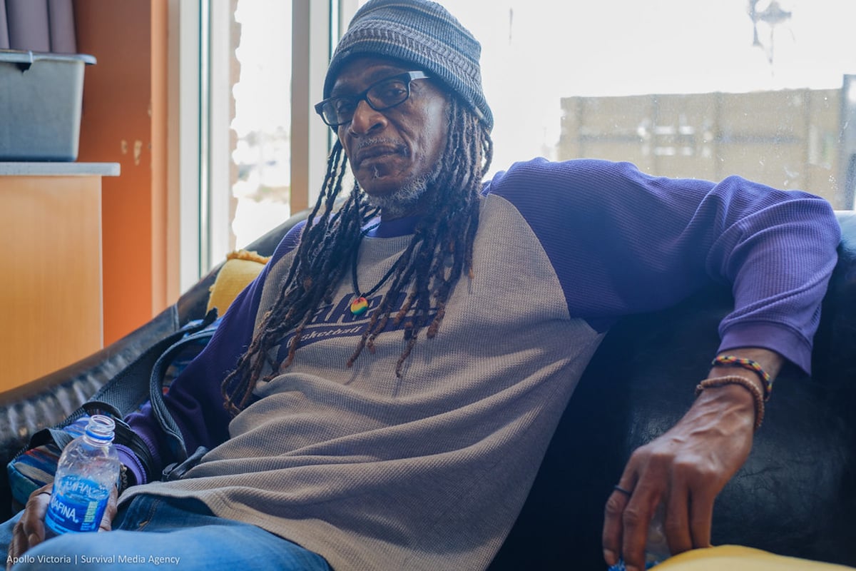 Brian Bukle wears a gray beanie cap and a t-shirt with purple sleeves, and leans back against a black couch he is sitting on.