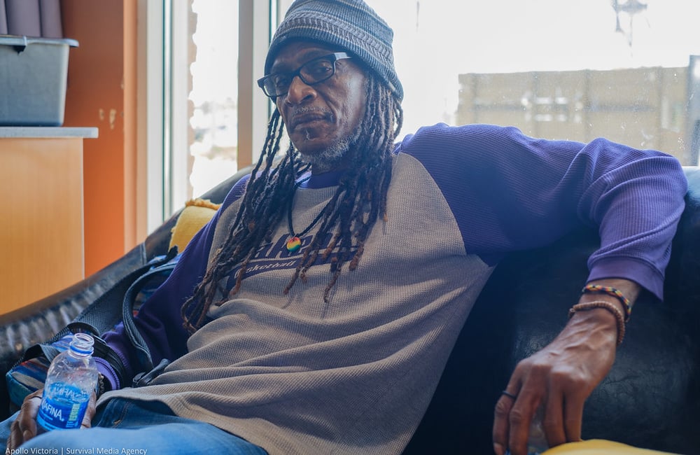 Brian Bukle wears a gray beanie cap and a t-shirt with purple sleeves, and leans back against a black couch he is sitting on.