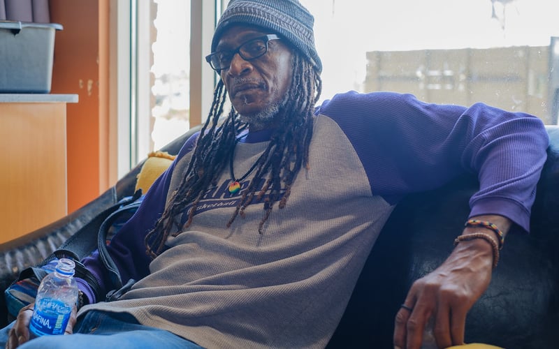 Brian Bukle wears a gray beanie cap and a t-shirt with purple sleeves, and leans back against a black couch he is sitting on.