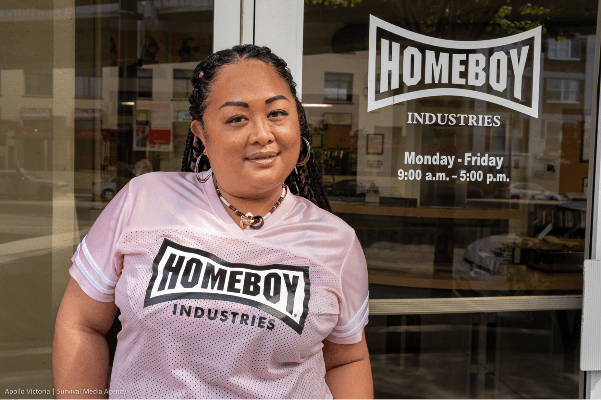 Maria wears a pink shirt and smiles in front of her place of work.