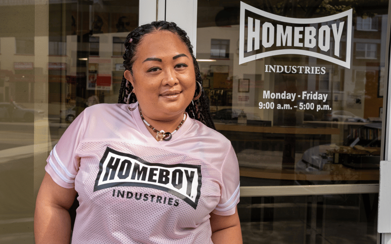 Maria wears a pink shirt and smiles in front of her place of work.