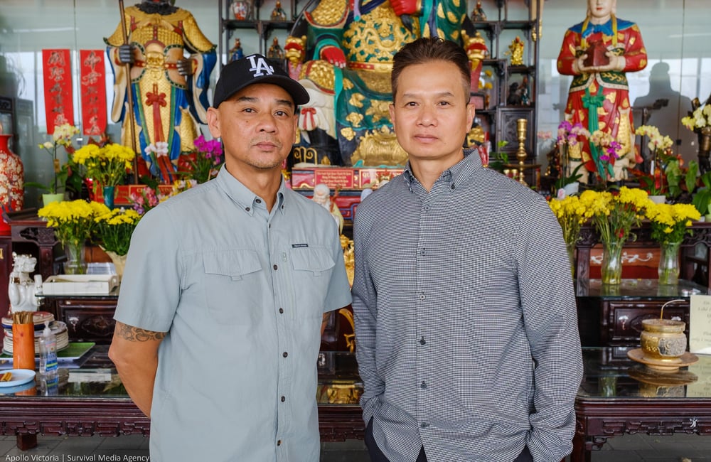 An and Tin pose for a photo in front of a large wooden altar laden with Buddhist figures, flowers, and incense. THey both wear button up shirts and look somber.