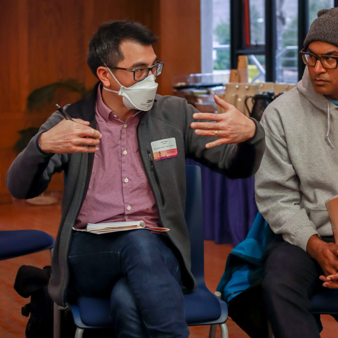 Carl Takei wears a face mask and nametag as he gestures while looking to his left. Another person sits beside Carl and looks at him.