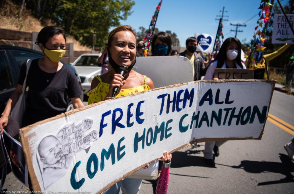 Ny Nourn stands in the street holding a banner reading "free them all come home Chanthon"