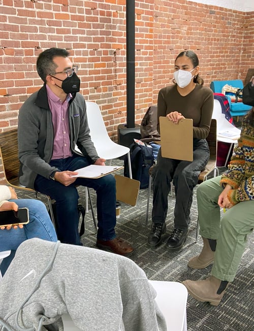A group of people wear face masks and sit in a circle as they have a discussion. Some have clipboards.