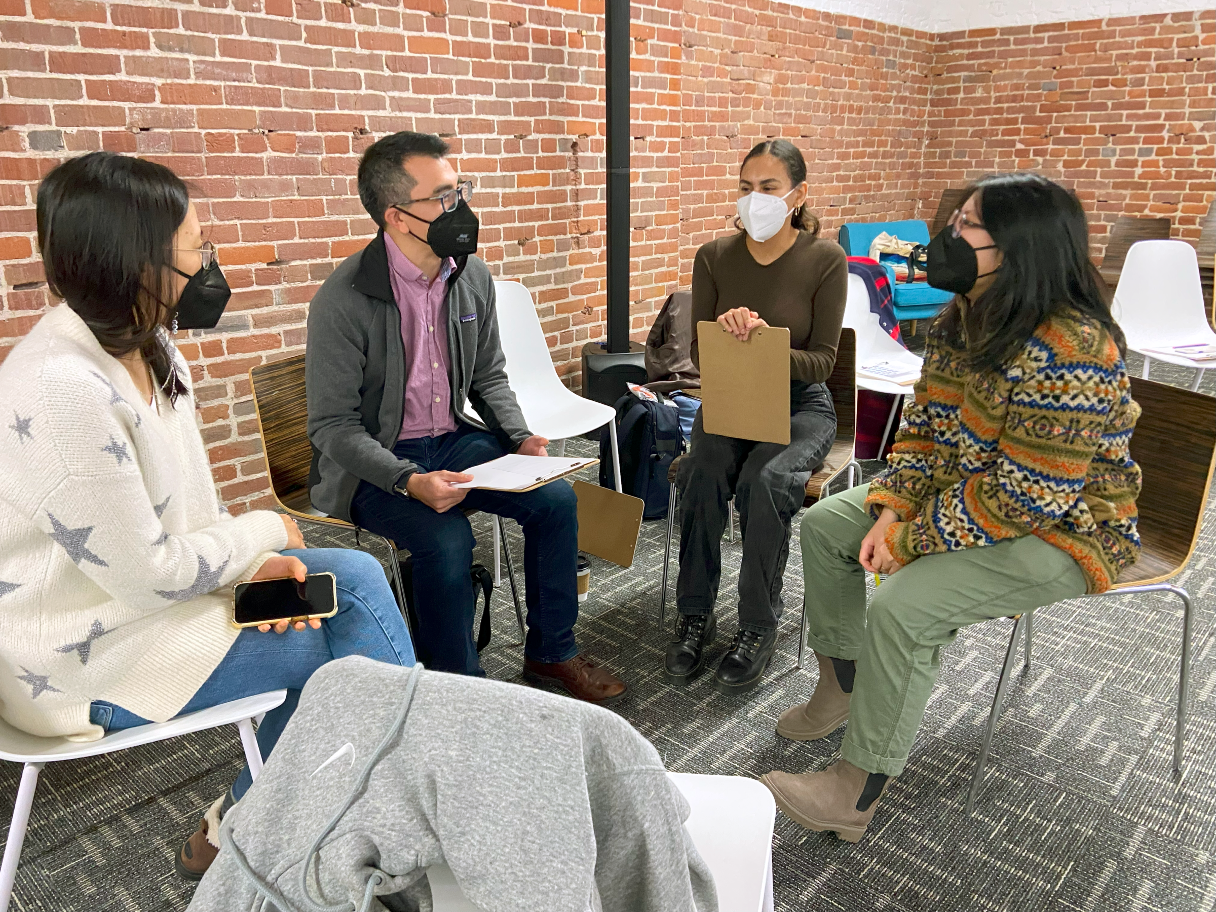 A group of people wear face masks and sit in a circle as they have a discussion. Some have clipboards.