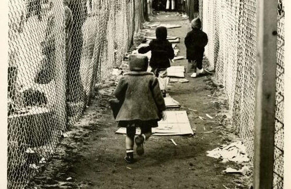 A black and white photo shows children in winter coats running a long a dirth path. Chain link fence lines both sides of the path. People stand against the fence in the foreground and at the end of the path in the background, where there is a large building. Visible on the ground are flat pieces of wood or cardboard.