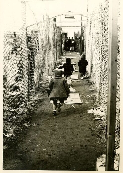 A black and white photo shows children in winter coats running a long a dirth path. Chain link fence lines both sides of the path. People stand against the fence in the foreground and at the end of the path in the background, where there is a large building. Visible on the ground are flat pieces of wood or cardboard.