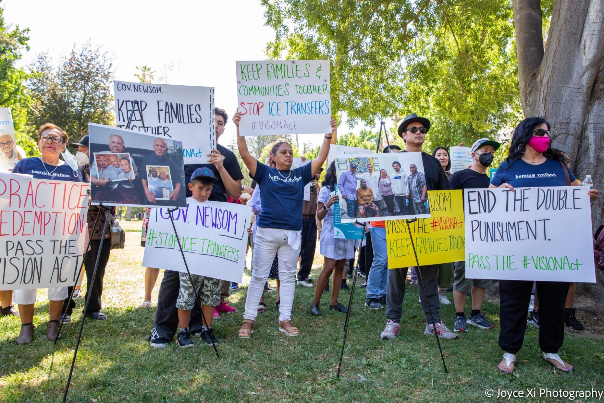 Community members hold up signs that read "keep families together" while they stand on a lawn in front of the CA state capitol.