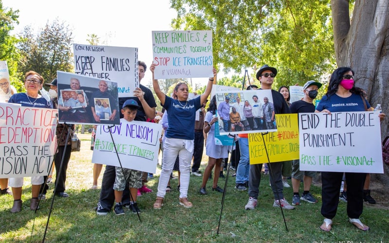 Community members hold up signs that read "keep families together" while they stand on a lawn in front of the CA state capitol.
