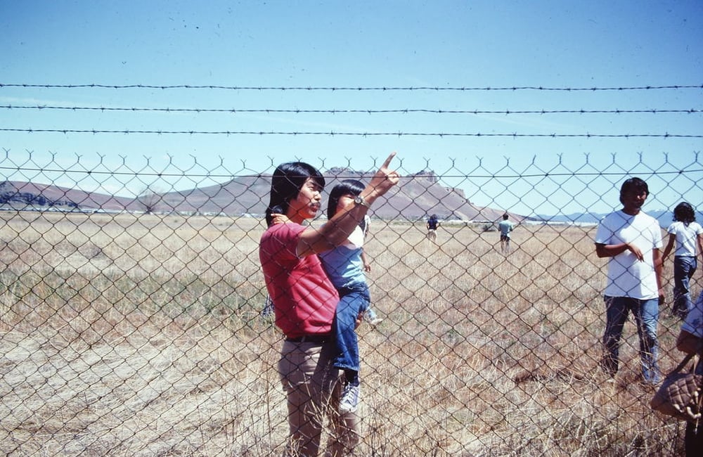 A man holds a child in one arm who looks as he touches the barbed wire fence in front of them. Dry grassland surrounds them. A little further behind is a group of other Japanese Americans participating in the Tule Lake pilgrimage. The hill called Castle Rock is in the background.