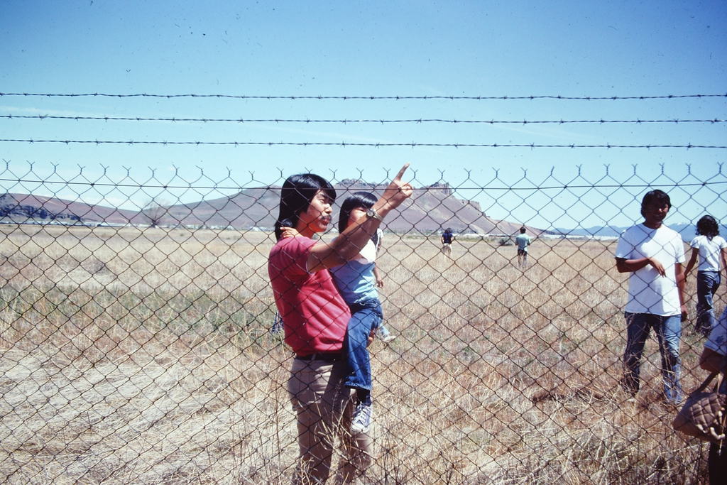 A man holds a child in one arm who looks as he touches the barbed wire fence in front of them. Dry grassland surrounds them. A little further behind is a group of other Japanese Americans participating in the Tule Lake pilgrimage. The hill called Castle Rock is in the background.