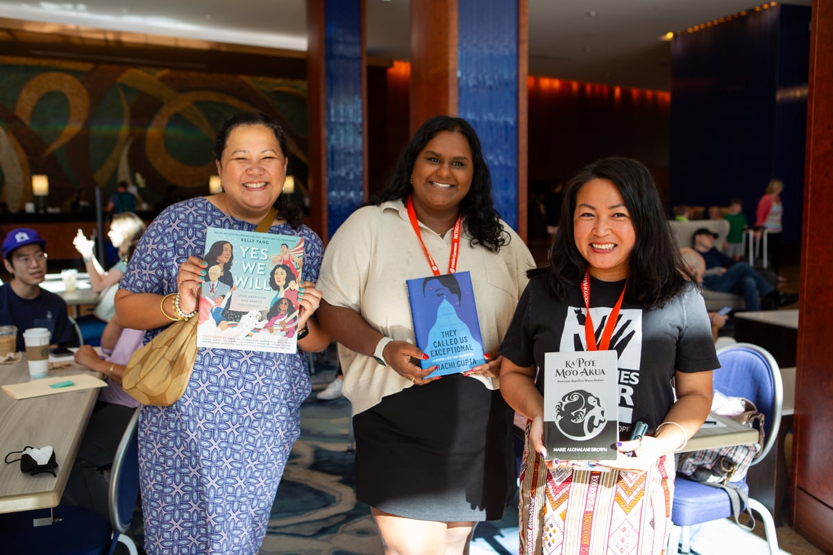 Three women smile as they hold up books for a photo. They wear orange lanyards and sunlight illuminates them inside a hotel conference room. One holds up "Yes We Will" by Kelly Yang, another person holds up "They Called Us Exceptional" by Prachi Gupta, and another holds up "Ka Po'e Mo'o Akua" by Marie Alohalani Brown.
