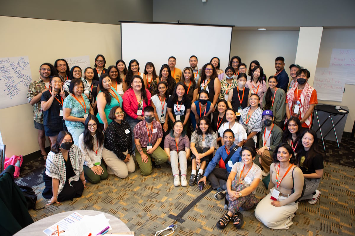 Over twenty people gather closely for a photo, some kneeling or sitting and some standing in front of a large projector screen with large post-it notes on the walls flanking them. Most wear nametags and orange lanyards.