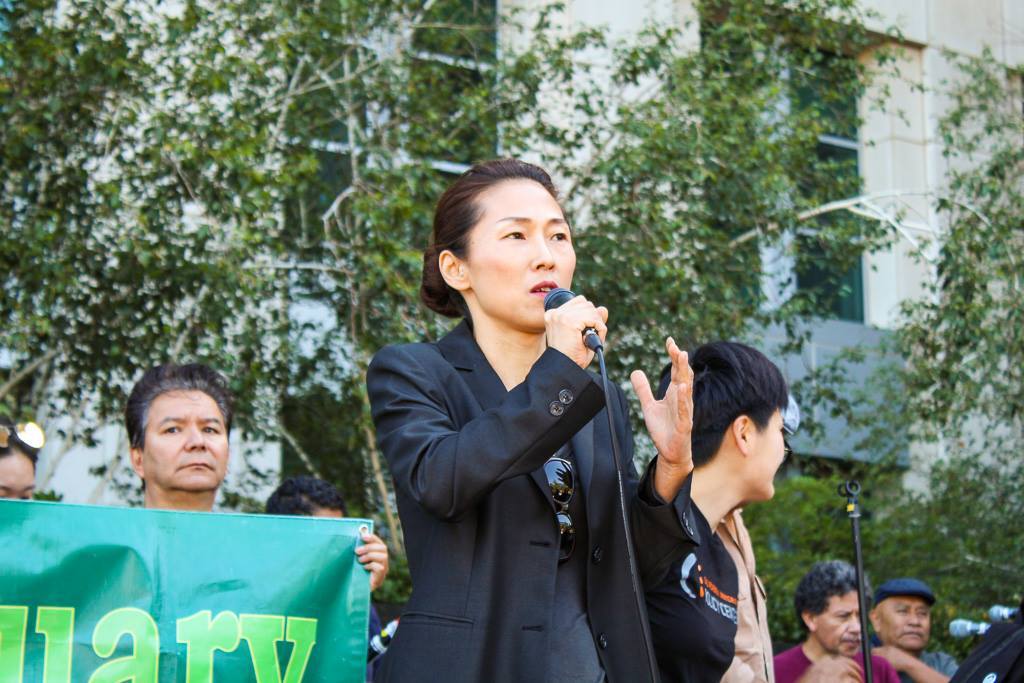 Woman in black jacket holds microphone with her right hand at a rally. Behind her, community members hold a green sign.