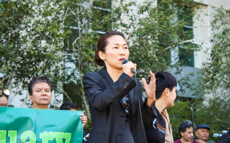 Woman in black jacket holds microphone with her right hand at a rally. Behind her, community members hold a green sign.
