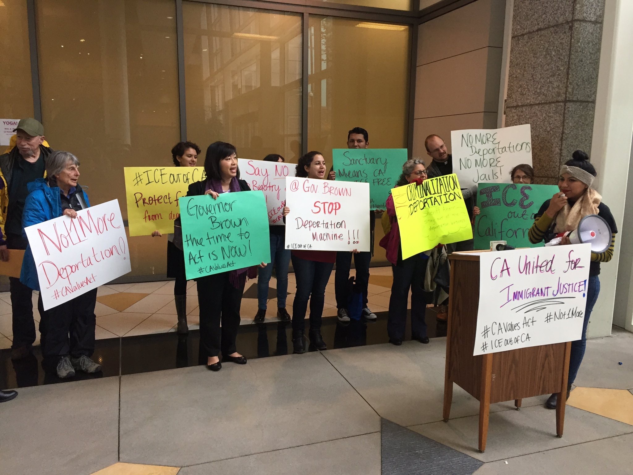 Nine people hold signs behind a podium with a sign on it reading "CA United for Immigrant Justice." A woman stands next to the podium holding a megaphone