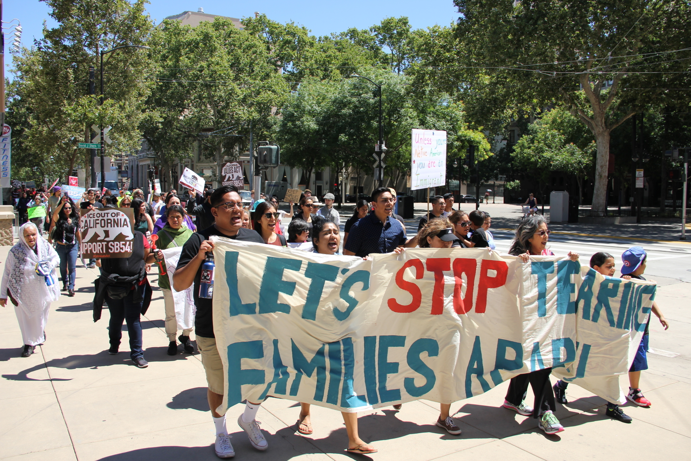 Protestors walk down the street holding a banner reading "let's stop tearing families apart"