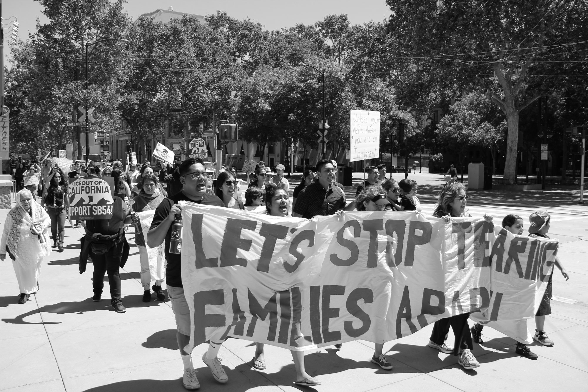 Black and white image of people holding banner reading "let's stop tearing families apart" followed by other protestors
