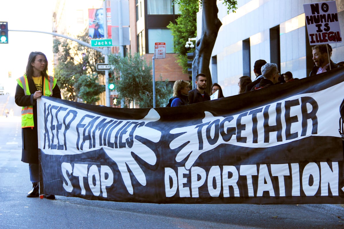 Person in yellow vest holds one side of banner reading "keep families together, stop deportation" that stretches onto a street.