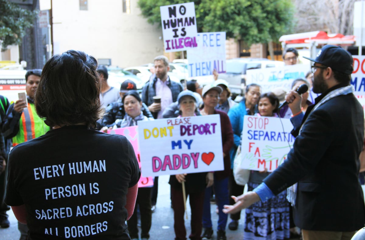 A crowd of people is gathered outside of the Department of Homeland Security office in San Francisco. They hold signs that say, "Free them all," "No human is illegal," and "Please don't deport my daddy."In the foreground a person wearing a patterned scarf gestures as he speaks into a microphone. A person with their back to the camera wears a shirt that says, "Every human person is sacred across all borders."