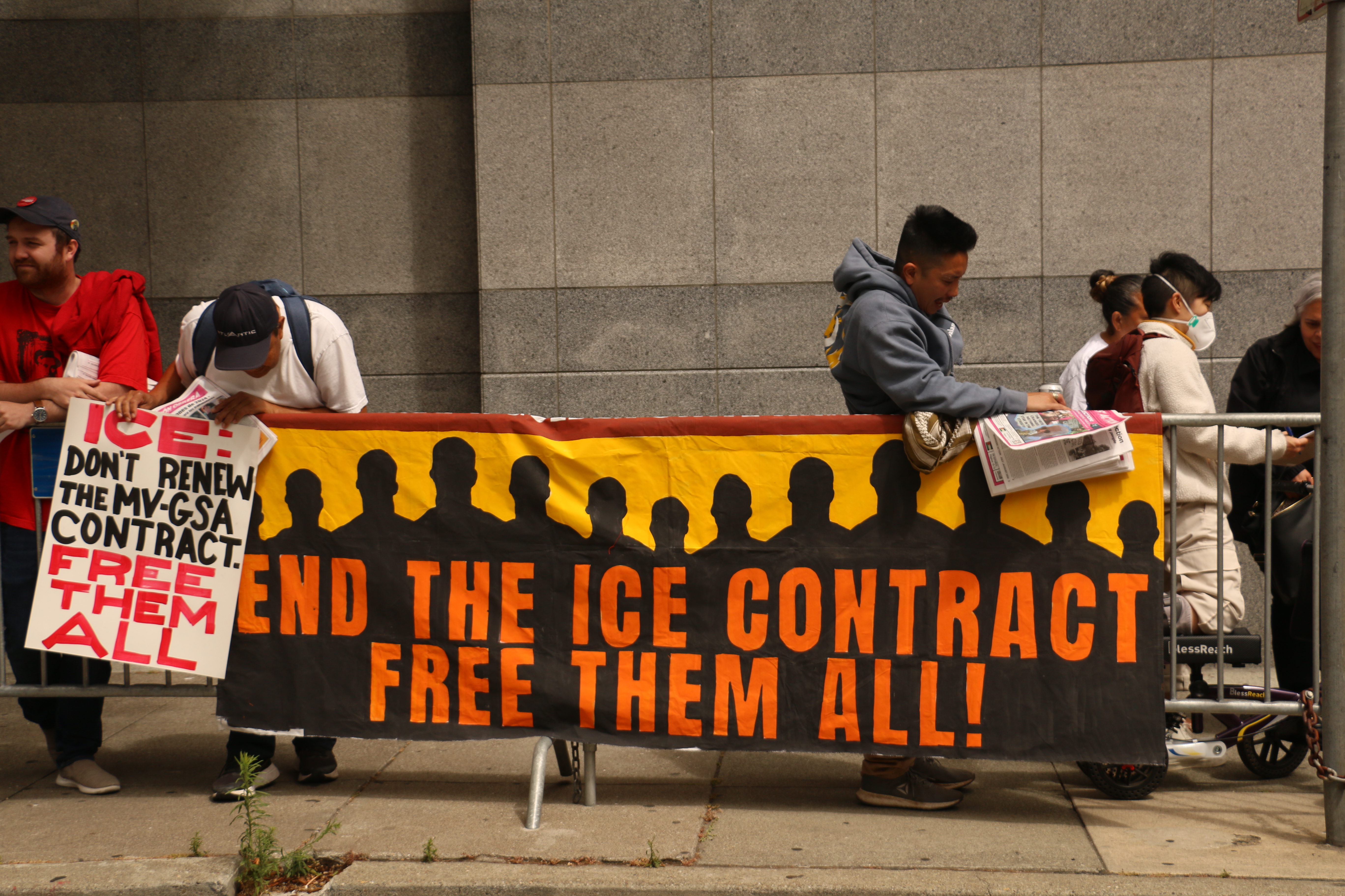 Supporters of the Mesa Verde and Golden State Annex strikes line the barricades on the sidewalk outside the San Francisco ICE Field Office. Draped over the barricade is a painted banner with the silhouettes of several people and the demand, "End the ICE Contract / Free them all!" One person leans over the barricade to view the banner. They hold their own painted sign that says, "ICE! Don't renew the MV-GSA Contract / Free them all."